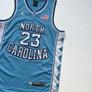 May include: Light blue basketball jersey with the words "NORTH CAROLINA" and the number "23" in white. The jersey has a V-neck, a small American flag patch, and the Jordan logo. The jersey is a size L.