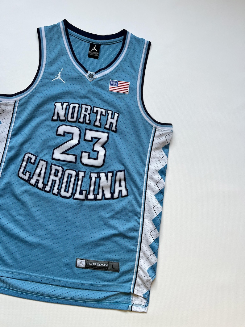 May include: Light blue basketball jersey with the words "NORTH CAROLINA" and the number "23" in white. The jersey has a V-neck, a small American flag patch, and the Jordan logo. The jersey is a size L.