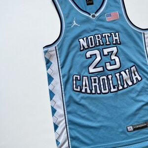 May include: Light blue basketball jersey with the words "NORTH CAROLINA" and the number "23" in white. Features a small American flag patch and the Jordan logo. The jersey has white and navy blue trim.