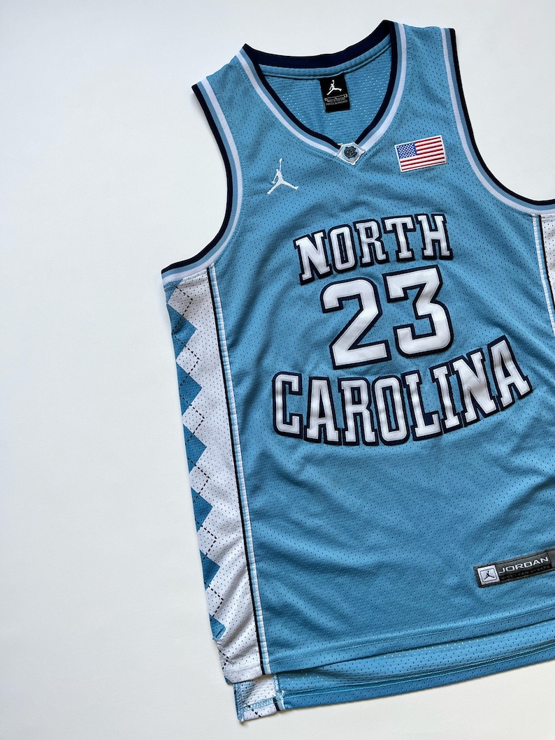 May include: Light blue basketball jersey with the words "NORTH CAROLINA" and the number "23" in white. Features a small American flag patch and the Jordan logo. The jersey has white and navy blue trim.
