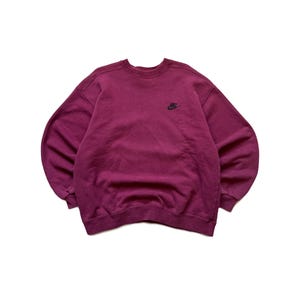 Puede incluir: Sudadera Nike de color granate con un peque&ntilde;o logotipo negro de Nike en la parte superior izquierda del pecho. La sudadera de cuello redondo est&aacute; hecha de un material suave y tiene mangas largas. La prenda se muestra sobre un fondo blanco.