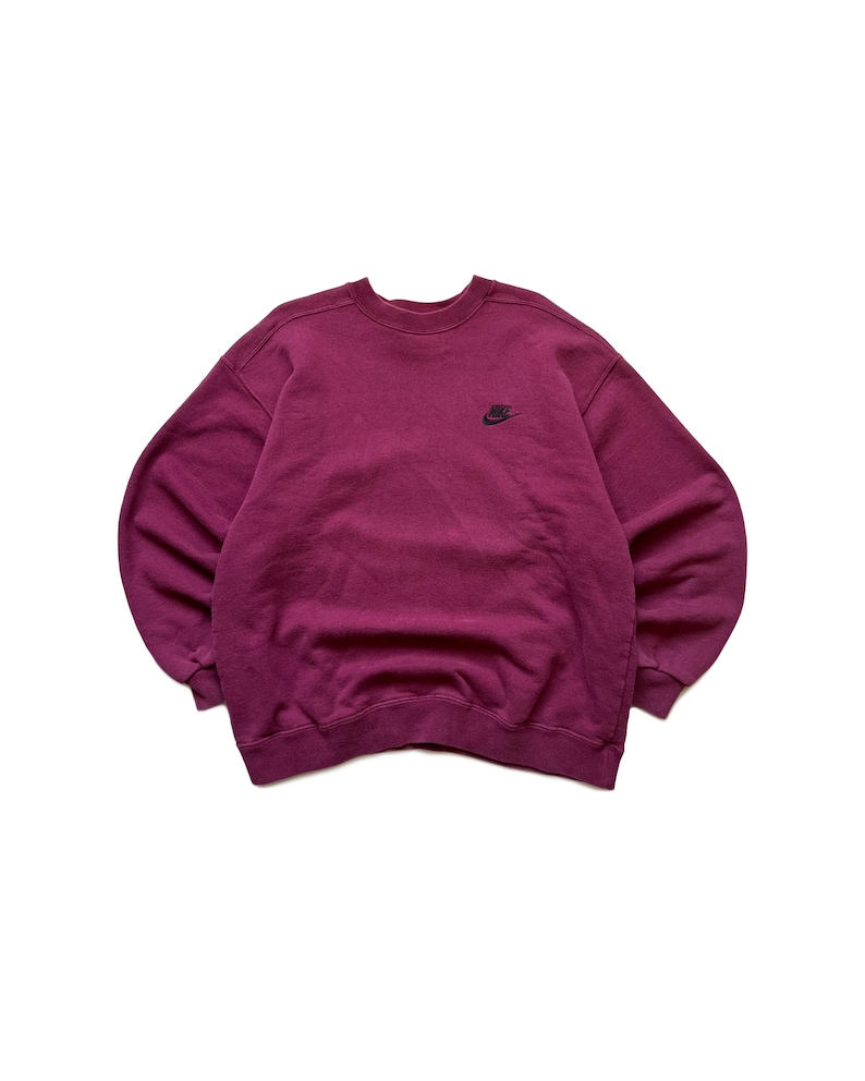 Puede incluir: Sudadera Nike de color granate con un peque&ntilde;o logotipo negro de Nike en la parte superior izquierda del pecho. La sudadera de cuello redondo est&aacute; hecha de un material suave y tiene mangas largas. La prenda se muestra sobre un fondo blanco.