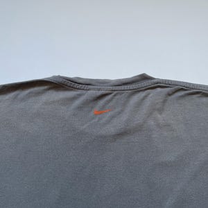 Puede incluir: Una camiseta gris de cuello redondo con un peque&ntilde;o logotipo naranja de Nike centrado en la parte superior de la espalda. La camiseta tiene un cuello acanalado y un aspecto ligeramente texturizado.
