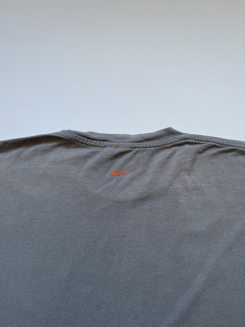 Puede incluir: Una camiseta gris de cuello redondo con un peque&ntilde;o logotipo naranja de Nike centrado en la parte superior de la espalda. La camiseta tiene un cuello acanalado y un aspecto ligeramente texturizado.