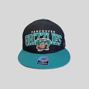 Può includere: Cappellino snapback dei Vancouver Grizzlies nero e verde acqua. Il cappellino presenta il nome della squadra in verde acqua e bianco, con un logo dell'orso grizzly. La visiera è verde acqua e ha una chiusura snapback.