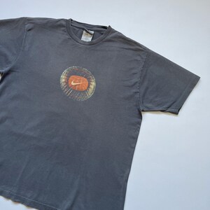 Puede incluir: Camiseta gris con un dise&ntilde;o gr&aacute;fico circular con el logotipo naranja de Nike. El gr&aacute;fico tiene un dise&ntilde;o texturizado y en capas con tonos naranja y marr&oacute;n. La camiseta est&aacute; hecha de un material suave y tiene un cuello redondo cl&aacute;sico.