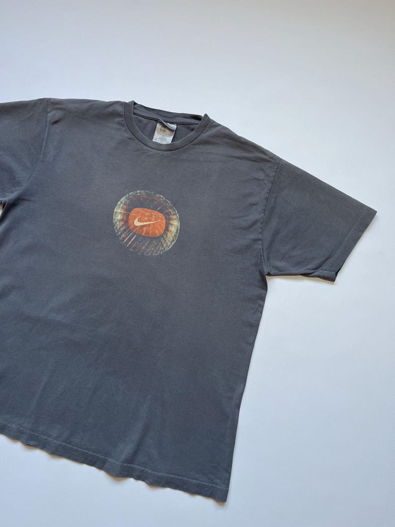 Puede incluir: Camiseta gris con un dise&ntilde;o gr&aacute;fico circular con el logotipo naranja de Nike. El gr&aacute;fico tiene un dise&ntilde;o texturizado y en capas con tonos naranja y marr&oacute;n. La camiseta est&aacute; hecha de un material suave y tiene un cuello redondo cl&aacute;sico.