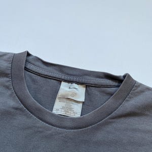 Puede incluir: Primer plano de una camiseta gris de cuello redondo con una etiqueta. La etiqueta presenta el logotipo de Nike e informaci&oacute;n sobre la talla. La camiseta es de color liso y parece estar hecha de un material suave.