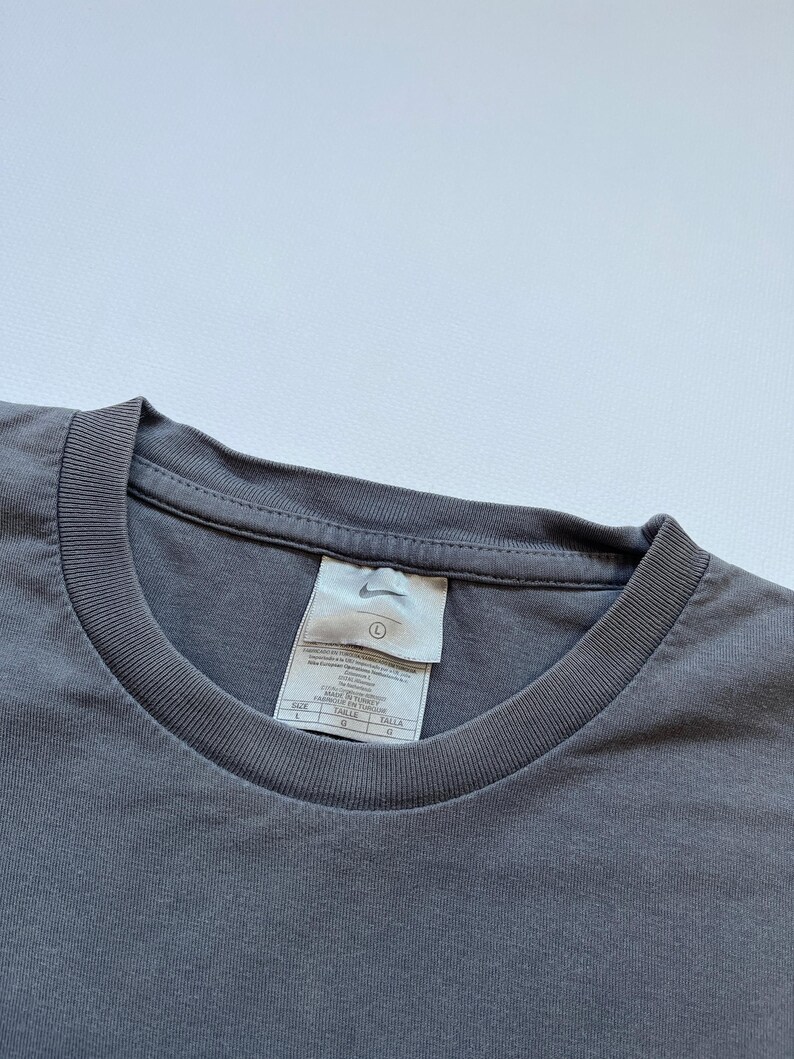 Puede incluir: Primer plano de una camiseta gris de cuello redondo con una etiqueta. La etiqueta presenta el logotipo de Nike e informaci&oacute;n sobre la talla. La camiseta es de color liso y parece estar hecha de un material suave.