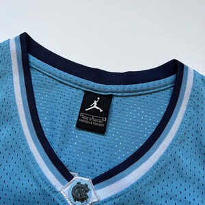 May include: Close-up of a light blue basketball jersey with a V-neck collar. The jersey features a white and navy blue striped trim, a patch with a logo, and a small American flag patch. The tag reads "Made in Thailand".