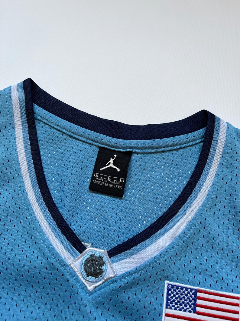 May include: Close-up of a light blue basketball jersey with a V-neck collar. The jersey features a white and navy blue striped trim, a patch with a logo, and a small American flag patch. The tag reads "Made in Thailand".