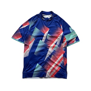 Puede incluir: Maillot deportivo azul con un patrón geométrico en rojo, blanco y turquesa. El maillot de manga corta tiene cremallera y un pequeño logotipo en el cuello. Diseñado para actividades deportivas.