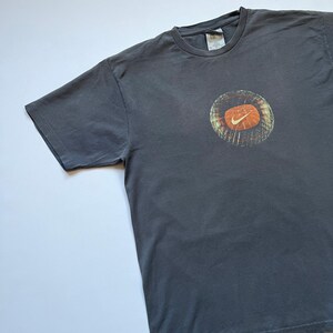 Puede incluir: Camiseta gris oscuro con un gr&aacute;fico circular con el logotipo de Nike naranja y blanco. La camiseta tiene cuello redondo y mangas cortas. El gr&aacute;fico tiene un aspecto vintage.