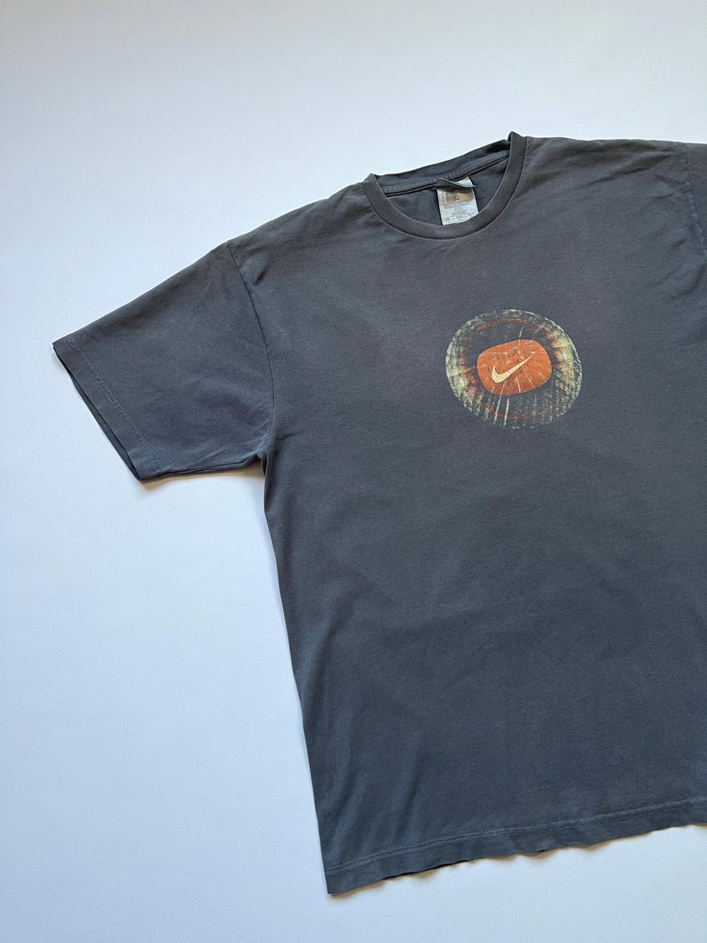 Puede incluir: Camiseta gris oscuro con un gr&aacute;fico circular con el logotipo de Nike naranja y blanco. La camiseta tiene cuello redondo y mangas cortas. El gr&aacute;fico tiene un aspecto vintage.