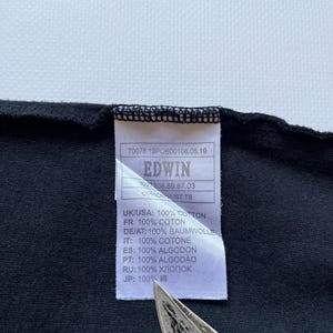K&ouml;nnte beinhalten: Nahaufnahme eines schwarzen Kleidungsst&uuml;cketiketts mit dem Markennamen "EDWIN" in Wei&szlig; aufgedruckt. Das Etikett enth&auml;lt auch den Text "100% COTTON" auf Englisch, Franz&ouml;sisch, Deutsch, Italienisch, Spanisch, Portugiesisch, Russisch und Japanisch.