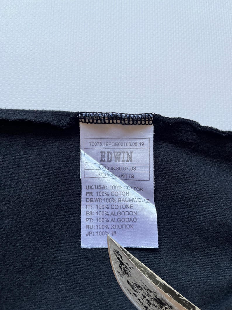 K&ouml;nnte beinhalten: Nahaufnahme eines schwarzen Kleidungsst&uuml;cketiketts mit dem Markennamen "EDWIN" in Wei&szlig; aufgedruckt. Das Etikett enth&auml;lt auch den Text "100% COTTON" auf Englisch, Franz&ouml;sisch, Deutsch, Italienisch, Spanisch, Portugiesisch, Russisch und Japanisch.