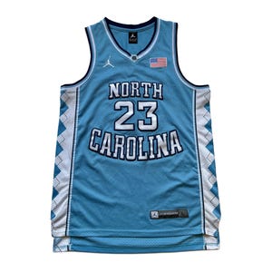 May include: Light blue North Carolina Tar Heels basketball jersey with white and navy blue accents. Features the number 23 and the words "North Carolina" in white. Includes an American flag patch and the Jordan logo.