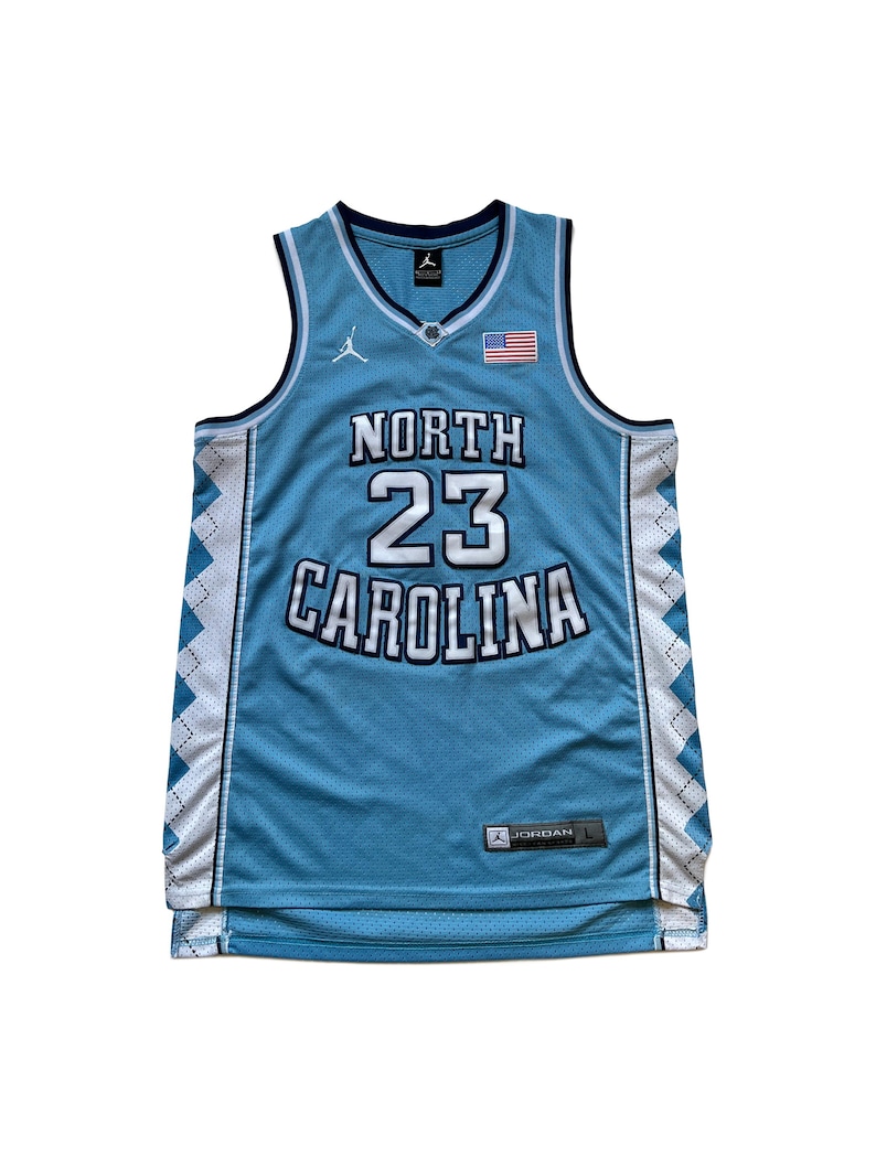 May include: Light blue North Carolina Tar Heels basketball jersey with white and navy blue accents. Features the number 23 and the words "North Carolina" in white. Includes an American flag patch and the Jordan logo.