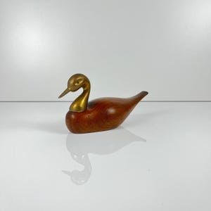 May include: A wooden duck figurine with a brass head and neck. The duck has a smooth, polished finish with a rich, reddish-brown color. The brass head and neck add a contrasting metallic accent to the wooden body. The duck is a decorative item.