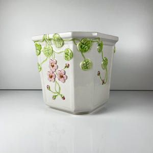 May include: White ceramic planter with a geometric shape, adorned with raised green leaves and pink flowers. The planter has a textured surface and is suitable for indoor or outdoor use.