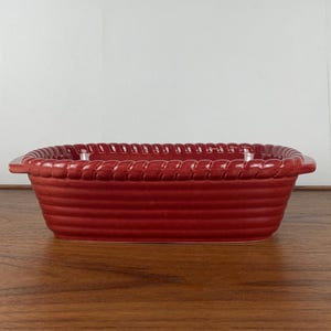 Op de afbeelding: Een rode keramische bakvorm met een touwachtig ontwerp rond de rand. De vorm heeft een rechthoekige vorm en is perfect voor het bakken van ovenschotels, brood of andere gerechten.