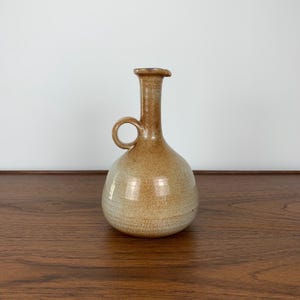May include: A brown ceramic jug with a rounded body and a small handle. The jug has a light brown glaze with a subtle white stripe.