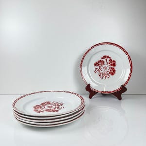 May include: White ceramic plates with a red floral design and scalloped red rims. One plate is displayed on a dark brown stand. A stack of four plates is visible in the foreground. The plates are on a white surface.