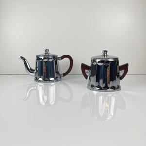 May include: A chrome-plated teapot and sugar bowl set. The teapot has a curved spout and a dark brown handle. The sugar bowl has two dark brown handles. Both pieces have a ridged design and a reflective surface.