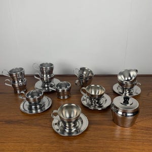 May include: A set of stainless steel teacups and saucers, including a sugar bowl with a lid. The cups and saucers are all shiny and reflect light. The set is arranged on a wooden surface.