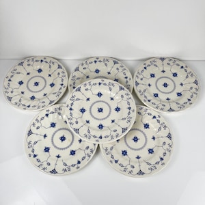 Peut inclure: Un ensemble de cinq assiettes rondes en céramique présentant une base de couleur crème avec un motif floral bleu répétitif. Chaque assiette possède un motif circulaire central entouré de délicats dessins de vignes et de bords festonnés.