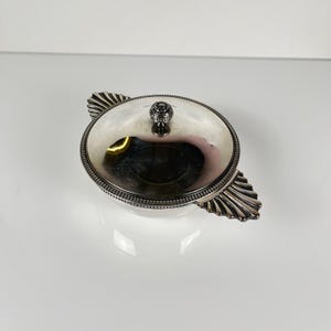 May include: A silver-colored, covered serving dish with a round shape and decorative handles. The lid features a small, ornate knob. The dish has a detailed, ridged border and is set against a white background.