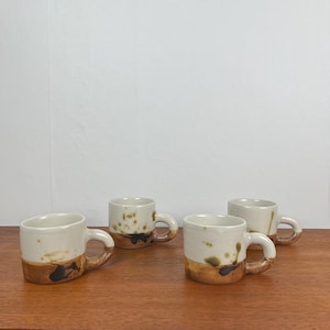 May include: Four ceramic mugs with a cream-colored upper half and a brown, textured lower half. The mugs have small handles and are arranged on a wooden surface. The mugs have a rustic, handmade appearance.