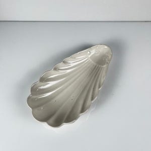 May include: A pale grey, shell-shaped dish with a scalloped edge. The dish has a glossy finish and a detailed, textured surface. The design is reminiscent of a clam shell, suitable for serving small items.