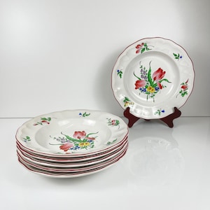 May include: A stack of white ceramic soup bowls with a red rim and floral design. The bowls have a scalloped edge and are decorated with red tulips, green leaves, and other colorful flowers. One bowl is displayed on a red stand.