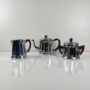 Könnte beinhalten: Ein Set aus drei verchromten Tee- oder Kaffee-Servierteilen. Das Set umfasst einen Sahnekännchen, eine Teekanne mit Ausguss und eine Zuckerdose mit Deckel. Jedes Teil hat einen dunkelbraunen Holzgriff und einen passenden Holzknopf auf dem Deckel.