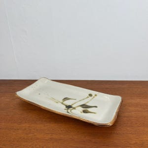 May include: A rectangular ceramic serving dish with a white glaze and a brown and green floral design. The dish has a gold rim.