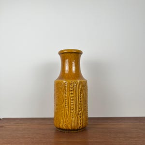 May include: A tall, mustard-yellow ceramic vase with a textured, vertical pattern. The vase has a rounded base, a cylindrical body, and a flared neck. The surface features a repeating design of vertical lines and small, raised dots.