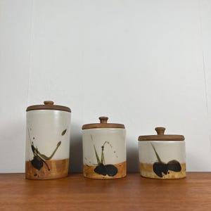May include: Three ceramic canisters with wooden lids. The canisters are white with brown and black abstract paint designs. The bottom of each canister has a brown band. The lids are round and made of wood.