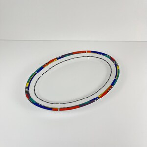 Può includere: Piatto ovale in ceramica bianca con un bordo geometrico colorato. Il bordo presenta un motivo ripetuto di rettangoli e linee rosse, gialle, blu e verdi. Una sottile linea nera corre lungo i bordi interno ed esterno.