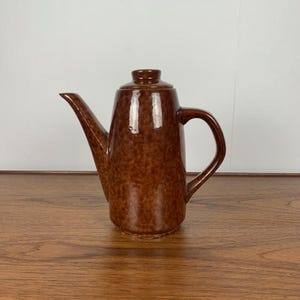 Peut inclure: Une théière en céramique brune avec un glaçage moucheté. La théière a un corps haut et mince et un bec verseur sur le côté. La poignée est du côté opposé au bec verseur.