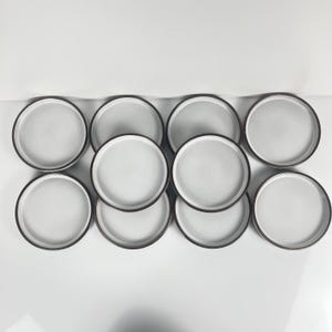 Peut inclure: Huit assiettes en céramique blanches avec un bord marron foncé. Les assiettes sont disposées en quinconce, mettant en valeur leur design simple et moderne. Les assiettes ont une surface légèrement texturée.