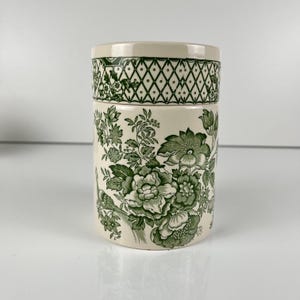 Peut inclure: Un pot cylindrique en céramique avec une base de couleur crème et un motif floral vert foncé. Le design présente des illustrations botaniques détaillées de grandes fleurs et de feuilles, avec une bordure en treillis en losange sur le couvercle.