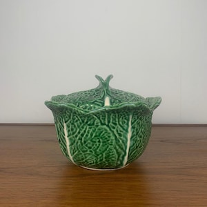 May include: A green ceramic cabbage-shaped serving dish with a lid. The dish is detailed with white veins and has a white interior.