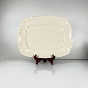 Peut inclure: Un plat de service rectangulaire blanc cassé avec un bord festonné. Il est soutenu par un support en bois brun foncé. La surface est lisse et réfléchissante, suggérant de la céramique ou de la porcelaine. Le design est classique et élégant.