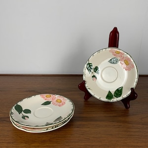 Peut inclure: Deux soucoupes en céramique blanche avec un motif floral représentant des roses roses et des feuilles vertes. Les soucoupes sont empilées l'une sur l'autre.