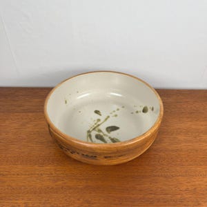 May include: A ceramic bowl with a light beige interior and a brown exterior. The bowl features a decorative design of green splatters and leaf-like shapes. The bowl is sitting on a wooden surface.