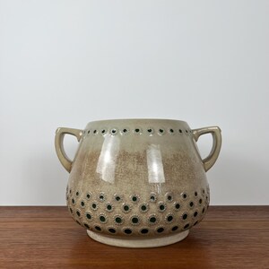 Peut inclure: Un vase en céramique beige avec deux anses. Le vase présente une surface texturée avec un motif de petits cercles vert foncé. Le bord supérieur comporte une rangée de petits trous. Le vase repose sur une petite base ronde.