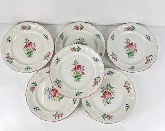May include: A set of five white ceramic plates with scalloped edges, each featuring a hand-painted floral motif in red, yellow, green, and blue. The plates have a thin red rim detail and are arranged in a cluster against a plain white background.