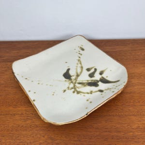 May include: Square ceramic plate with a cream-colored surface and a gold-colored rim. The plate features a dark green and brown abstract design. The plate is sitting on a wooden surface.