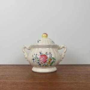 May include: A white ceramic sugar bowl with a lid, featuring a floral design in shades of pink, yellow, blue, and green. The bowl has two handles and a rounded shape.